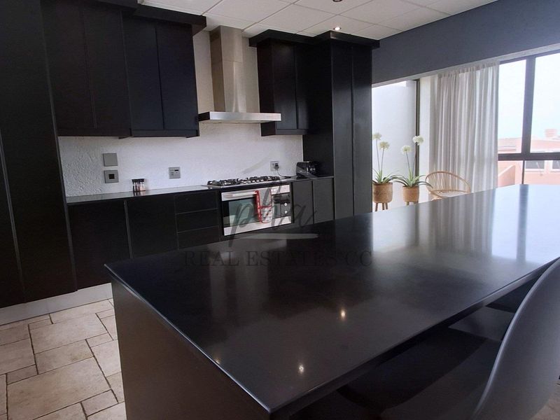 Modern kitchen featuring sleek black cabinetry, a large central island with stone countertop, stainless steel appliances, and tiled flooring with natural light.