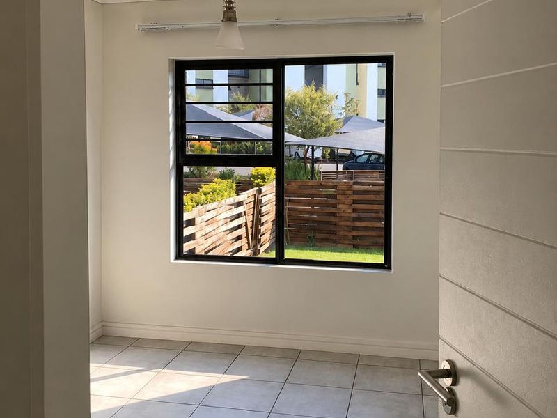A bright, empty bedroom featuring neutral tiled flooring, a modern pendant light, and a large window with security bars overlooking the complex grounds.
