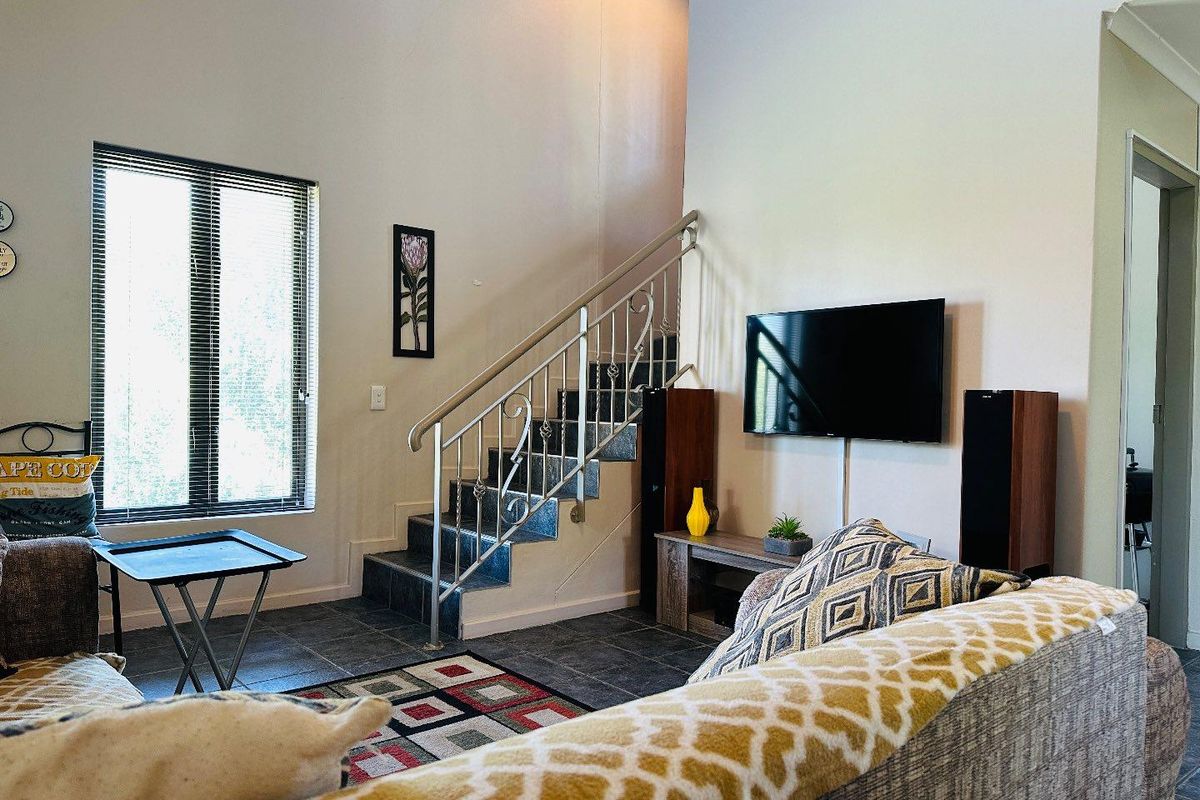 A living room featuring a staircase with a metal balustrade, tiled flooring, and large windows with blinds, offering a bright and open atmosphere.