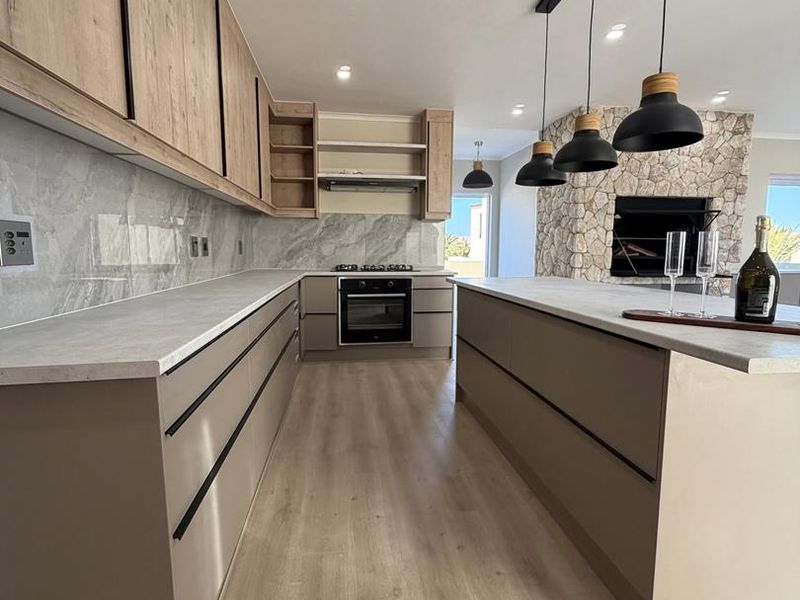 Modern open-plan kitchen featuring sleek cabinetry, stone-look backsplash, central island, and a striking stone fireplace, creating a seamless living and dining space.