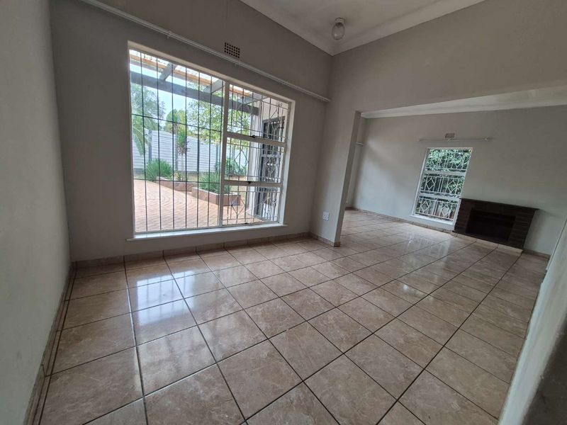 Spacious, light-filled living area featuring neutral tiled flooring, a classic brick fireplace, and large windows with security bars overlooking the rear garden.