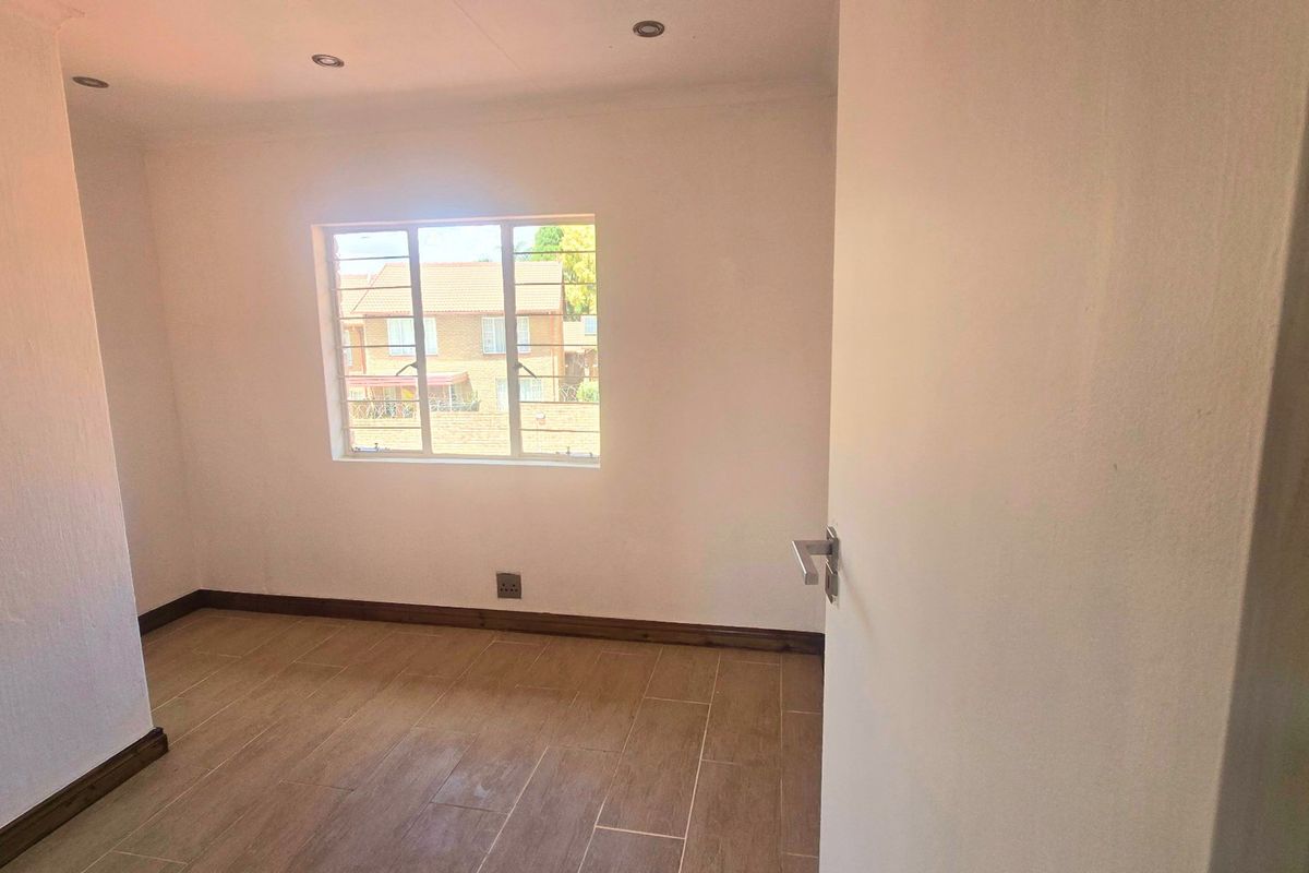 A bright, empty bedroom featuring modern wood-look tiled flooring, recessed downlights, and a window fitted with security burglar bars.