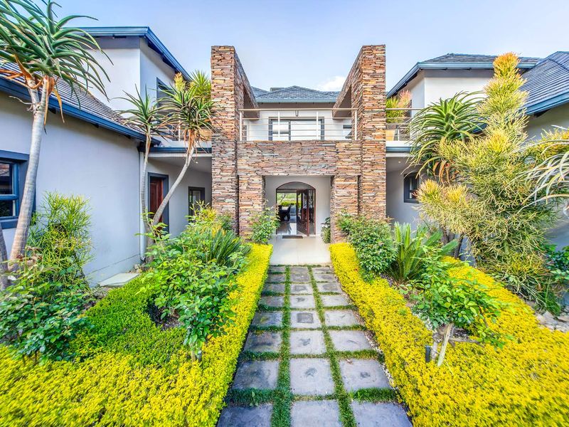 A striking front entrance featuring stone cladding, a paved walkway through a landscaped garden, and an upper-level balcony.
