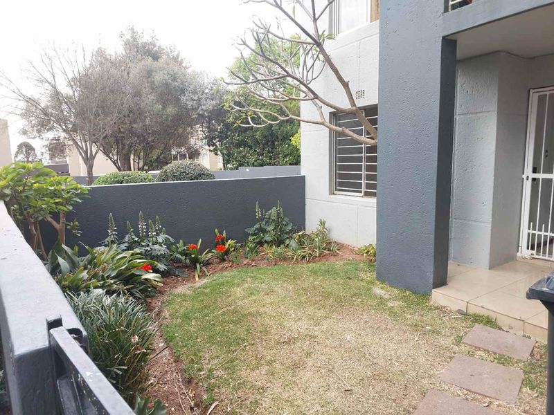 A private rear garden area featuring a small lawn, landscaped garden beds, a tiled patio section, and secure access to the apartment.