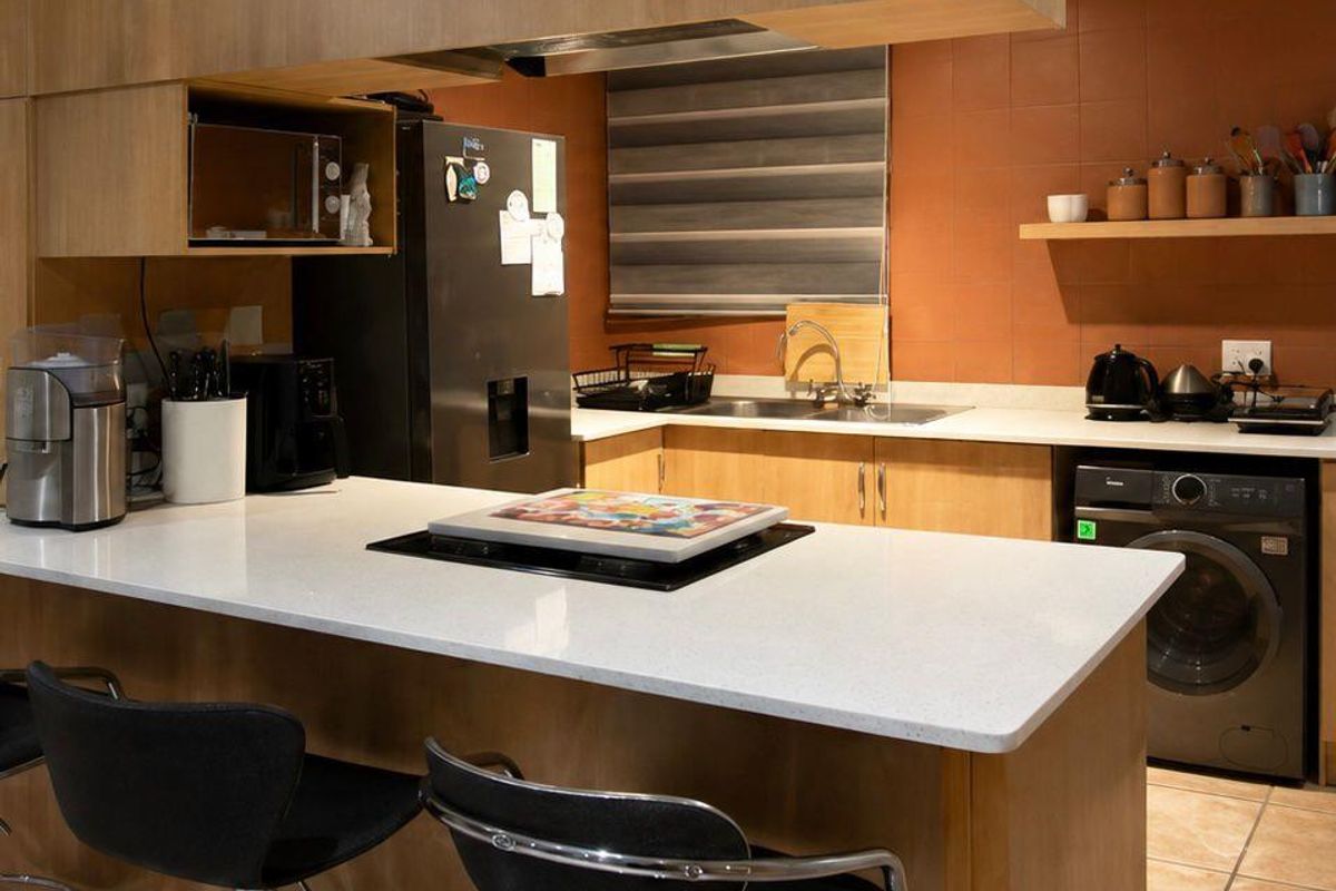A functional kitchen featuring light wood cabinetry, a white stone breakfast bar, integrated hob, and tiled splashbacks with an adjacent laundry area.
