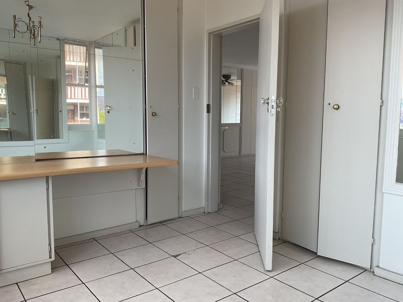 A vacant bedroom featuring built-in cupboards, a vanity area with mirror, and tiled flooring, offering a neutral canvas for potential updates.