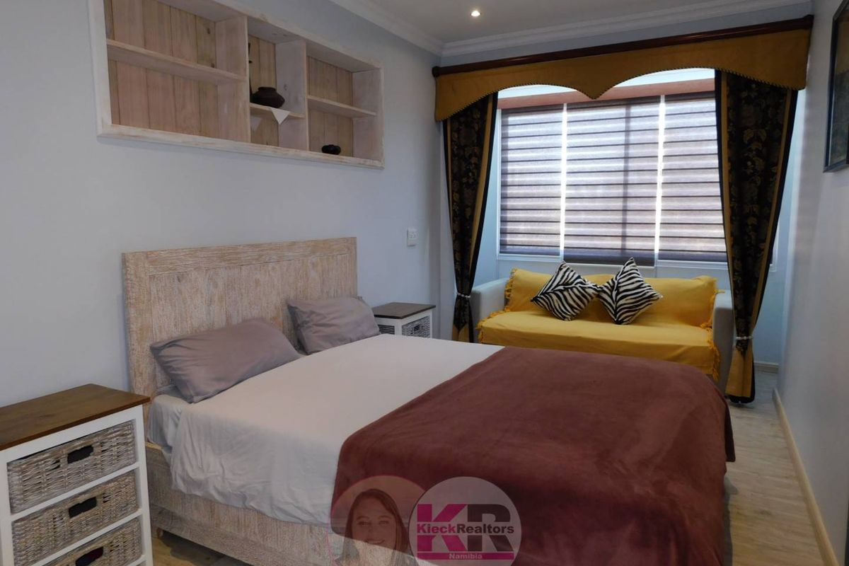 A well-maintained bedroom featuring built-in wooden shelving, recessed ceiling lighting, and large windows with fitted blinds.