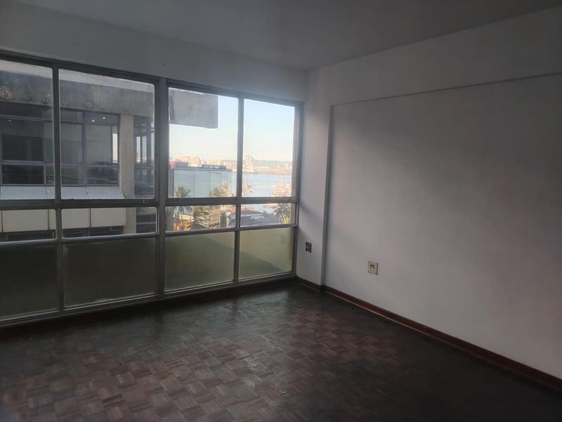 An empty living room featuring original parquet flooring and large windows offering an urban view, providing a blank canvas for interior design.