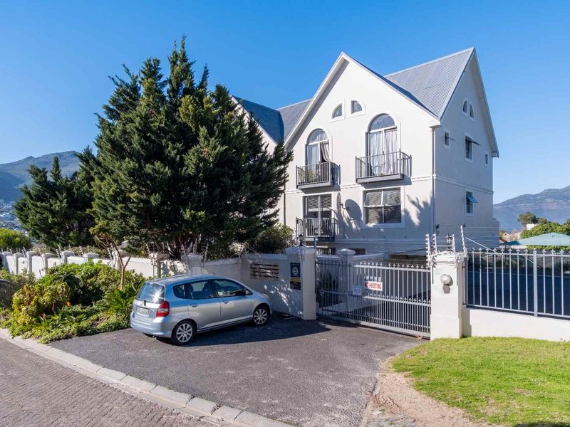 Front elevation of a multi-story apartment building featuring a secure gated entrance, paved driveway, and balconies with mountain views.