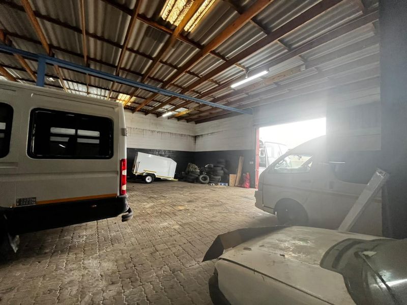 Spacious industrial garage featuring exposed timber roof trusses, corrugated metal roofing, paved flooring, and a large roller shutter door for vehicle access.