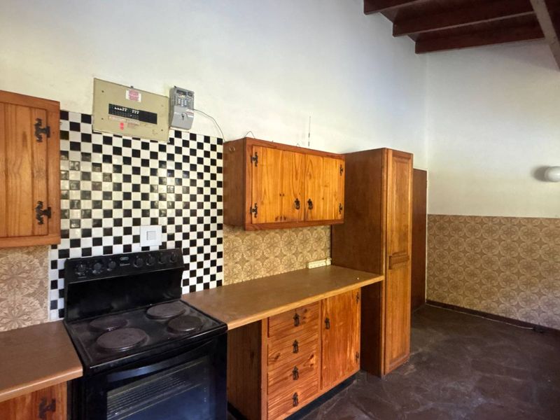 A dated kitchen featuring wooden cabinetry, a checkerboard tile backsplash, an electric stove, and exposed ceiling beams, offering significant potential for modernization.