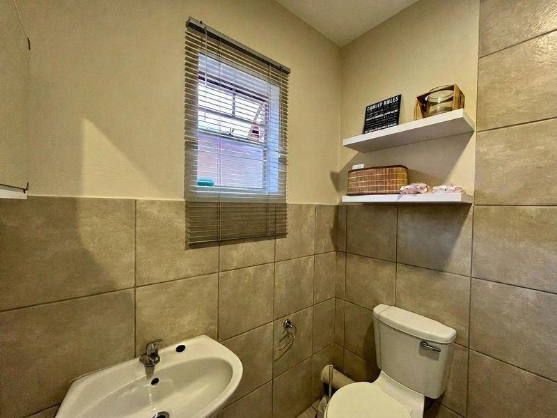 A compact bathroom featuring neutral ceramic wall tiles, a pedestal washbasin, a toilet, and convenient floating wall shelves.