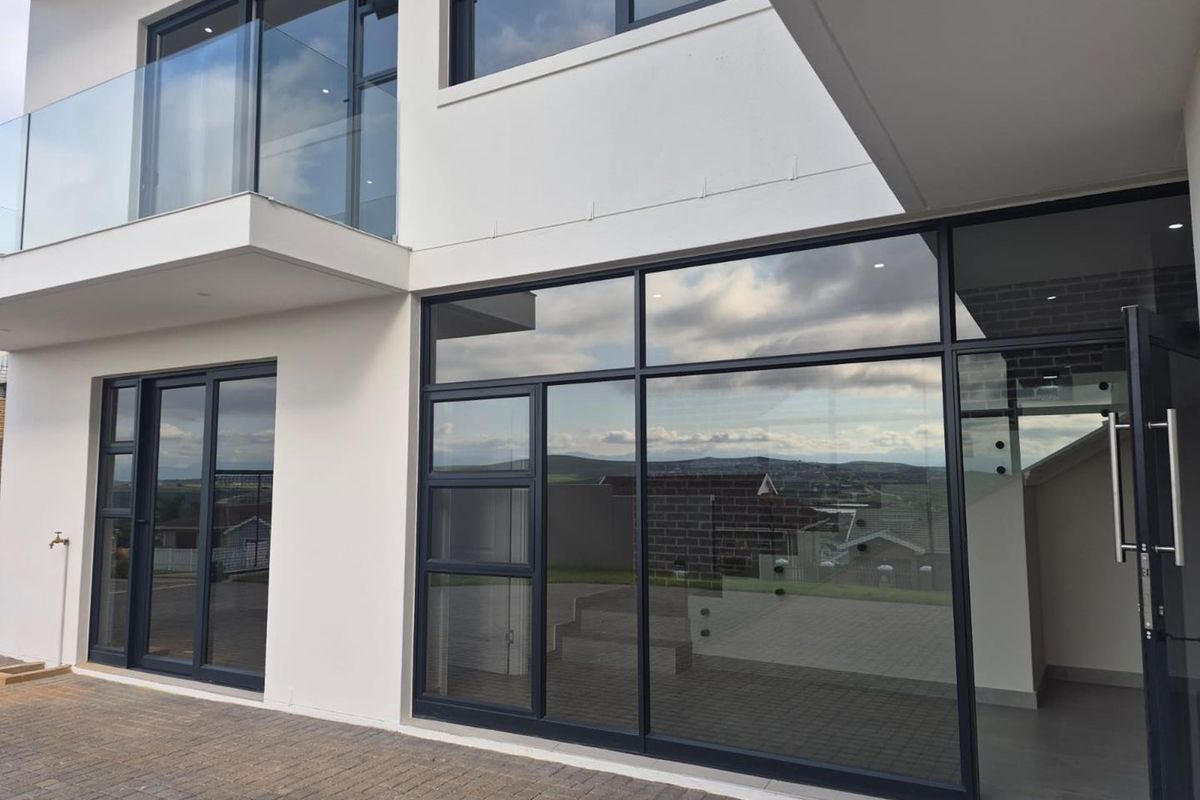 Modern rear exterior view featuring large aluminium-framed glass sliding doors, a paved patio area, and a glass balustrade on the upper balcony.