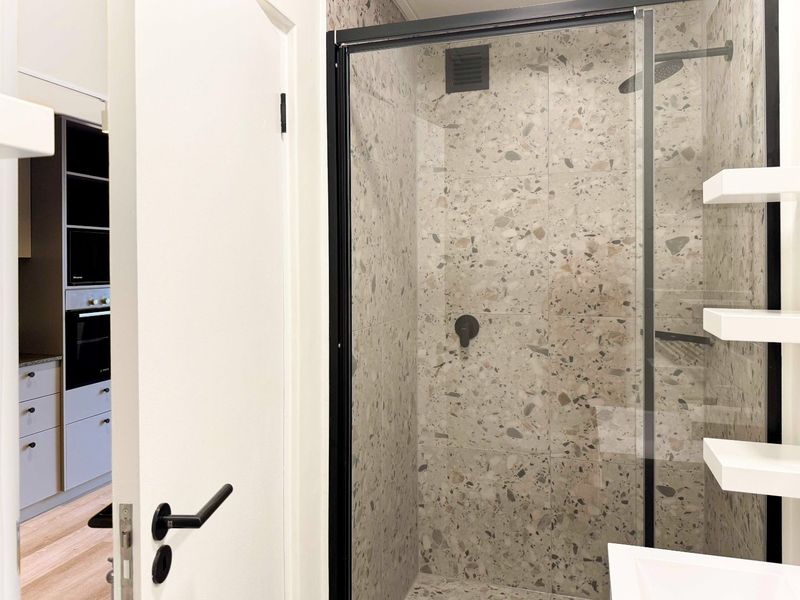 Modern bathroom featuring terrazzo-style tiling, a sleek black-framed glass shower enclosure, and built-in shelving, with a view into the adjacent kitchen area.