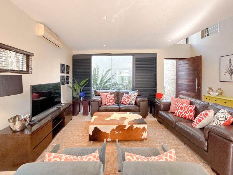 A well-maintained living room featuring tiled flooring, recessed lighting, a wall-mounted air conditioning unit, and large shutters opening to an outdoor area.