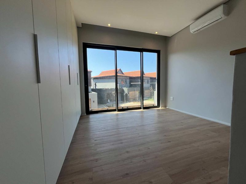A bright, modern bedroom featuring light wood-look laminate flooring, floor-to-ceiling built-in cupboards, a split-system air conditioner, and large sliding doors leading to a balcony.