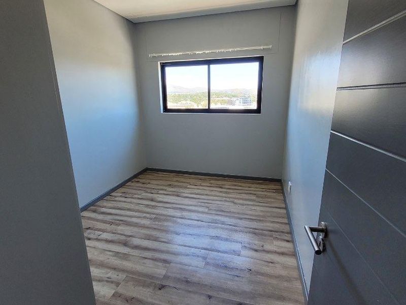 A bright, empty bedroom featuring modern laminate wood-look flooring, neutral grey walls, and a sliding window with natural light.