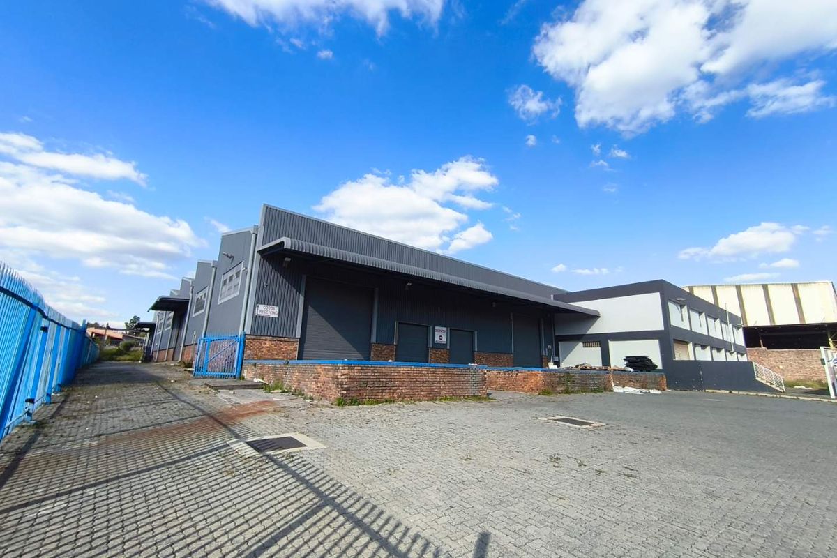 Industrial warehouse facility featuring multiple roller shutter doors, a raised loading dock, and a paved yard area enclosed by security fencing.