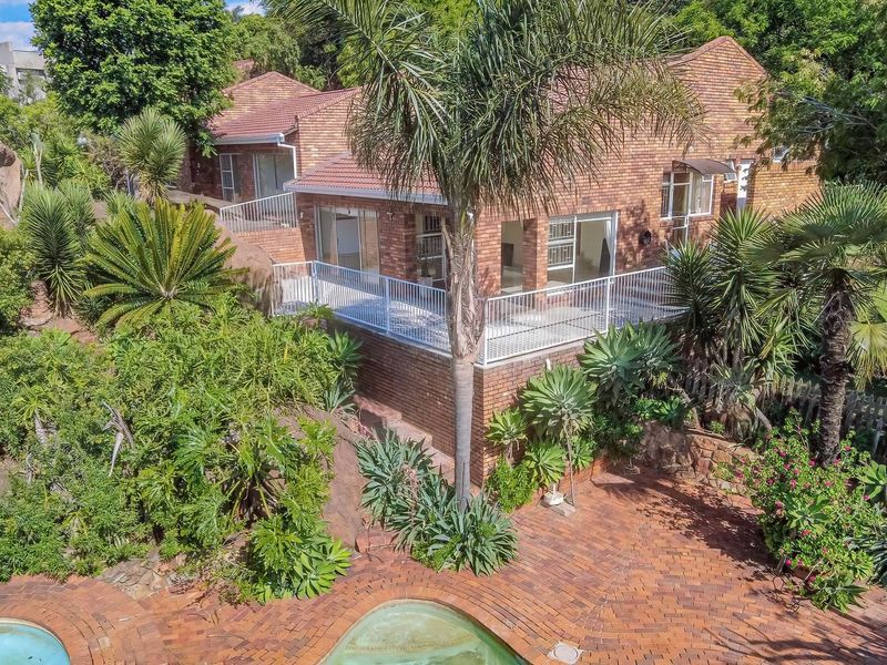 Elevated rear view of a brick house featuring a raised balcony, paved patio area, and a swimming pool set within a landscaped garden.