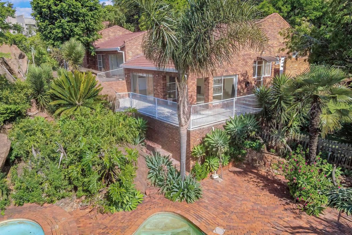 Elevated rear view of a brick house featuring a raised balcony, paved patio area, and a swimming pool set within a landscaped garden.