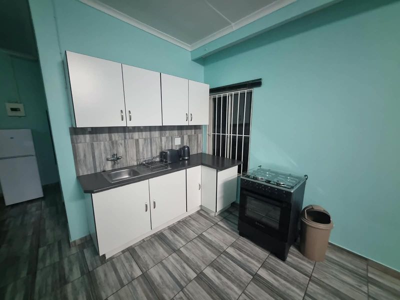 A functional kitchen featuring white cabinetry, dark laminate countertops, tiled backsplash, and tiled flooring, equipped with a freestanding stove and security bars on the window.