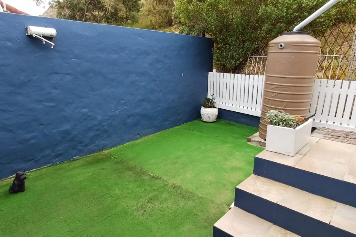 A compact rear courtyard featuring artificial turf, a blue boundary wall, a water storage tank, and tiled steps leading to the property.