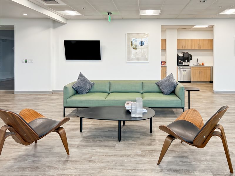 A modern open-plan office lounge area featuring a kitchenette with built-in cabinetry, recessed ceiling lighting, and durable wood-look laminate flooring.