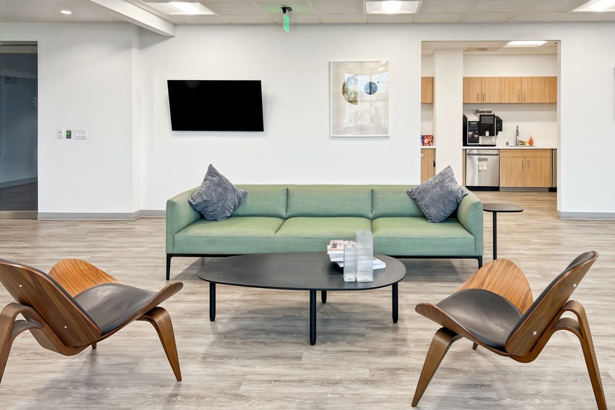 A modern open-plan office lounge area featuring a kitchenette with built-in cabinetry, recessed ceiling lighting, and durable wood-look laminate flooring.