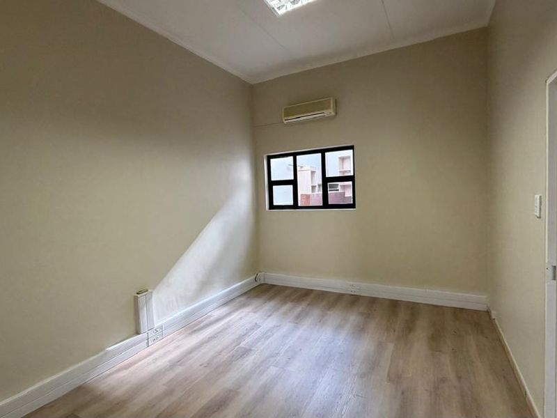 A clean, empty office space featuring light-toned laminate flooring, neutral walls, a wall-mounted air conditioning unit, and a window providing natural light.