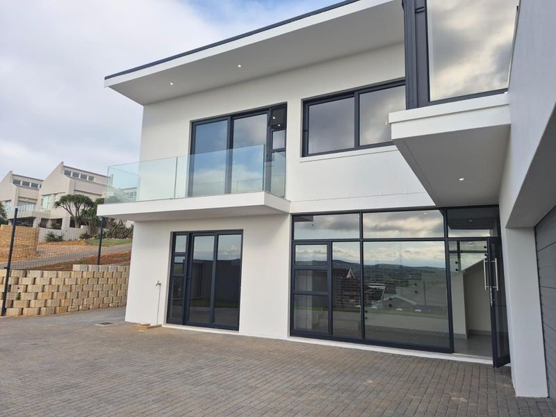 Modern double-storey residential facade featuring clean white finishes, large aluminium-framed windows, a glass balcony balustrade, and a paved driveway.