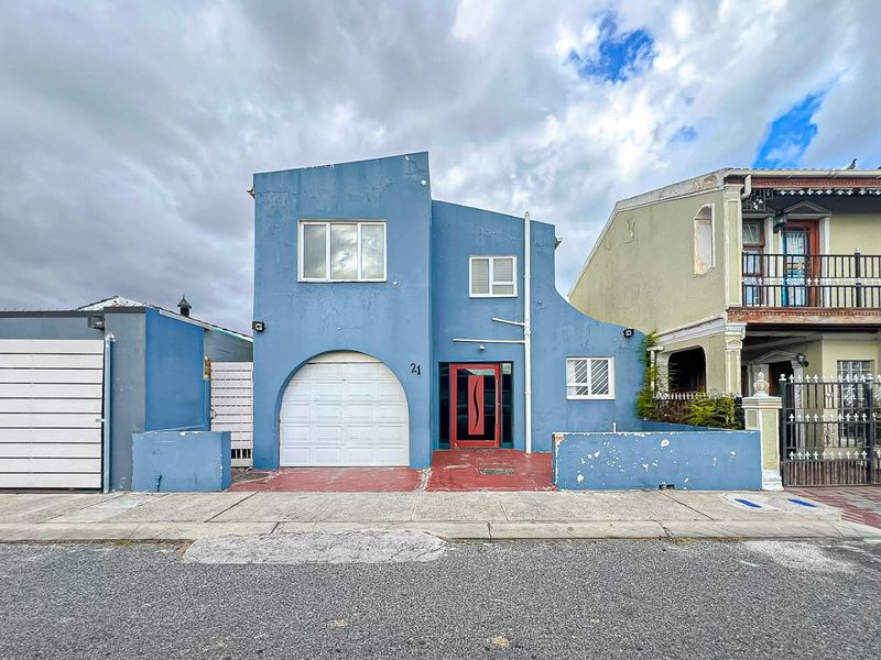 Front elevation of a blue house featuring a single garage, red front door, and a low boundary wall with visible wear to the exterior finishes.
