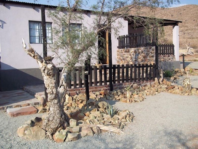 Front elevation of a rustic farm cottage featuring stone cladding, a covered veranda, and a timber picket fence set in a desert landscape.