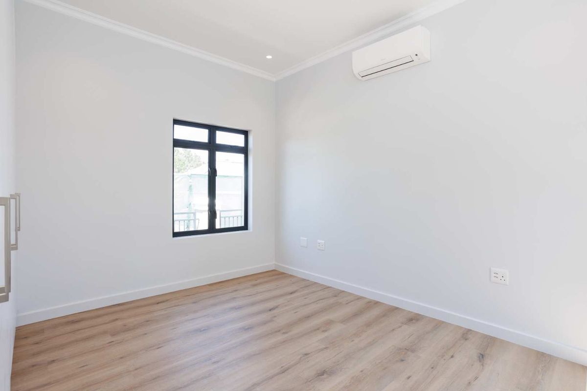 A bright, modern bedroom featuring light wood-look flooring, neutral walls, a black-framed window, and a wall-mounted air conditioning unit.