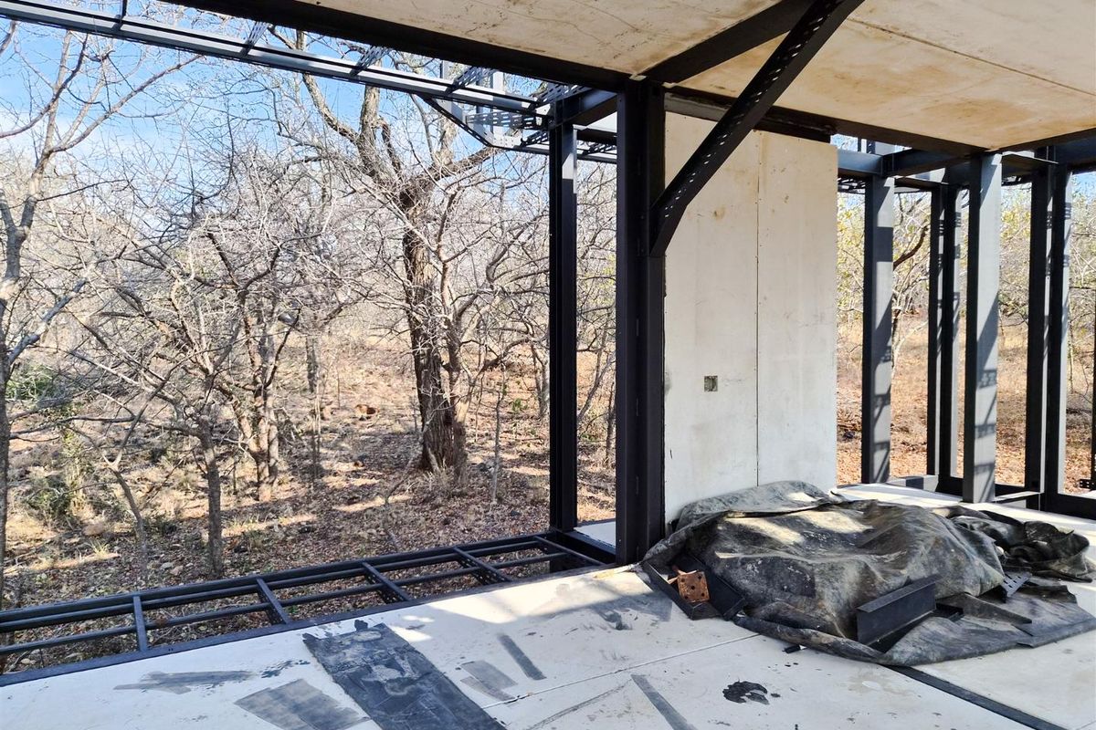 An unfinished interior space featuring an exposed steel structural frame and large windows overlooking a natural wooded landscape.
