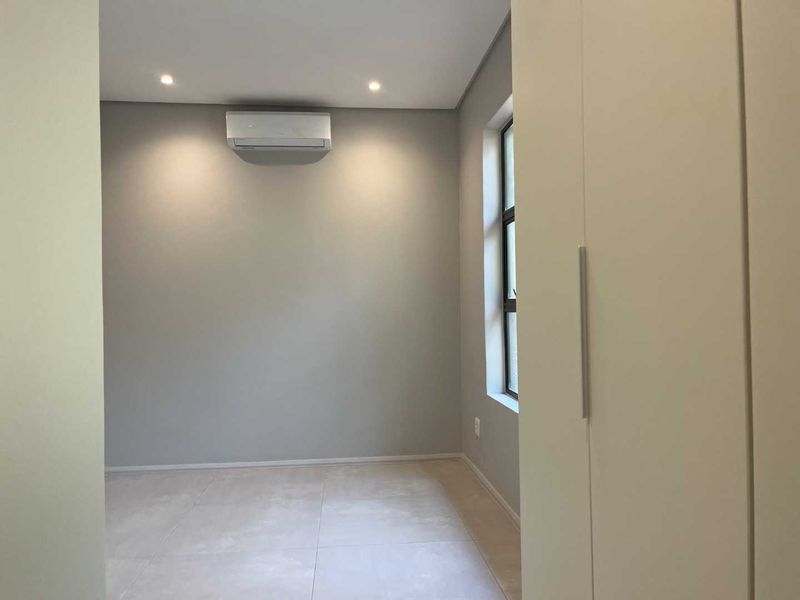 A clean, modern bedroom featuring neutral tiled flooring, recessed lighting, a built-in wardrobe, and a wall-mounted air conditioning unit.