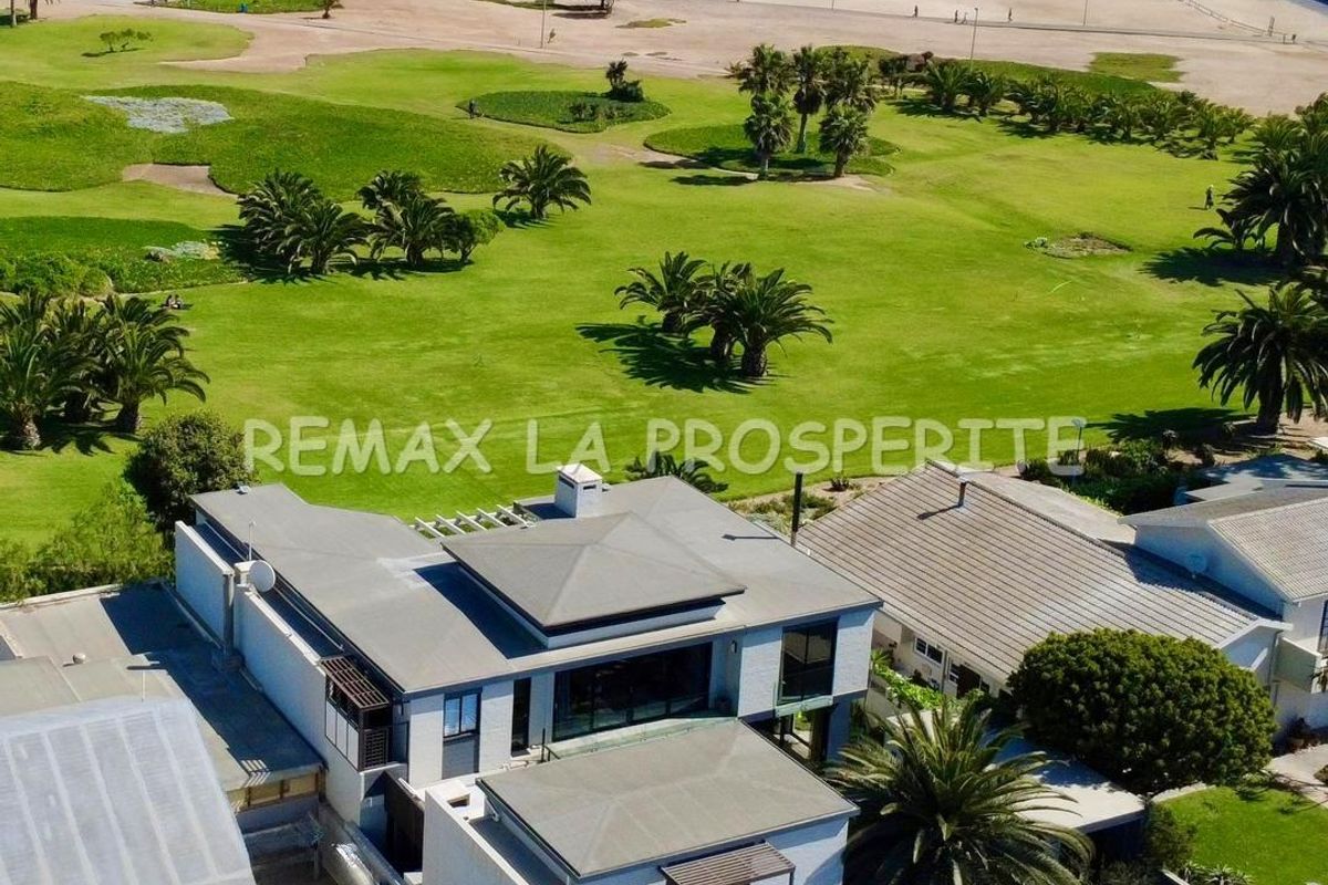 Aerial view of a modern multi-level coastal residence featuring flat roof architecture, balconies, and proximity to the beach and parkland.