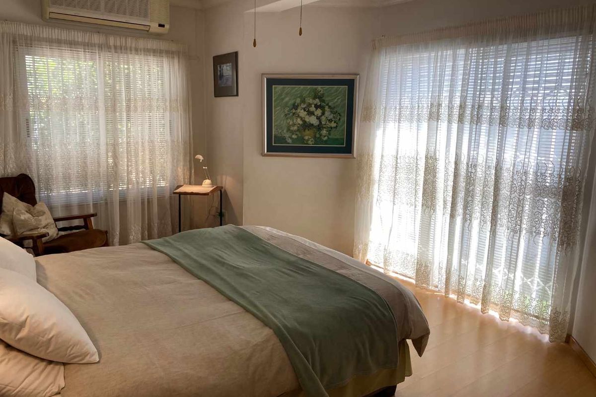 A well-lit bedroom featuring light-toned laminate flooring, large windows with sheer curtains, and a wall-mounted air conditioning unit.