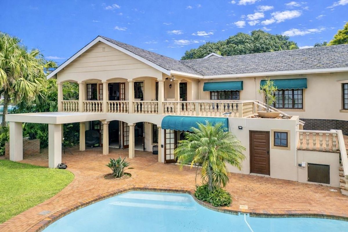 Spacious rear exterior featuring a swimming pool, brick-paved patio, covered balcony with balustrades, and established garden surroundings.