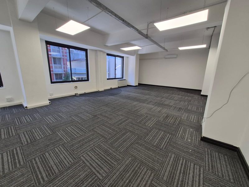 A spacious, open-plan office area featuring modern carpet tiles, suspended LED lighting, and large windows providing ample natural light.