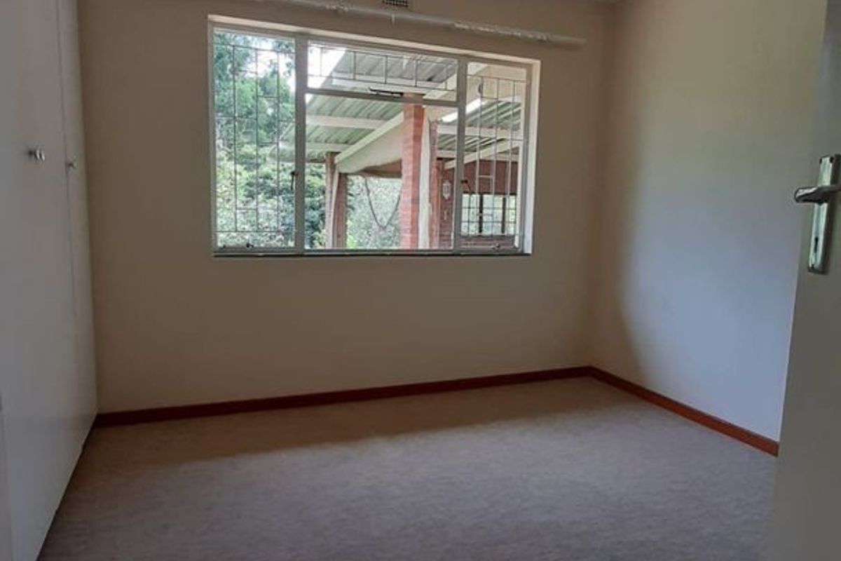 A neutral, empty bedroom featuring built-in cupboards, a ceiling fan, and a window with security bars overlooking the rear patio area.