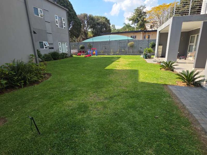 A well-maintained rear garden featuring a spacious lawn, paved patio area, and boundary wall, adjacent to the apartment building with balcony access.