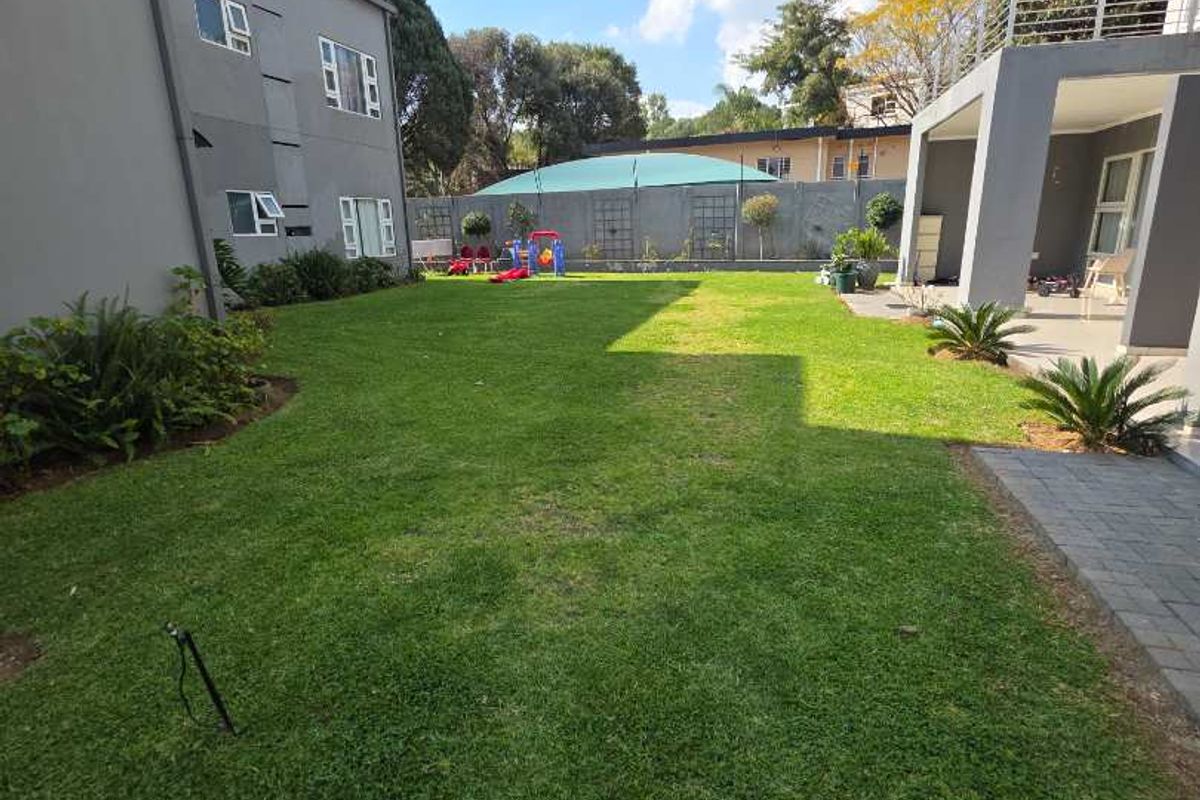A well-maintained rear garden featuring a spacious lawn, paved patio area, and boundary wall, adjacent to the apartment building with balcony access.