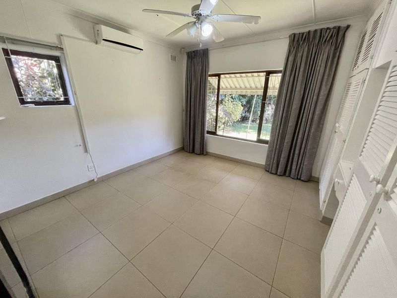 A bright, empty bedroom featuring tiled flooring, built-in cupboards, a ceiling fan, and a split-system air conditioner.