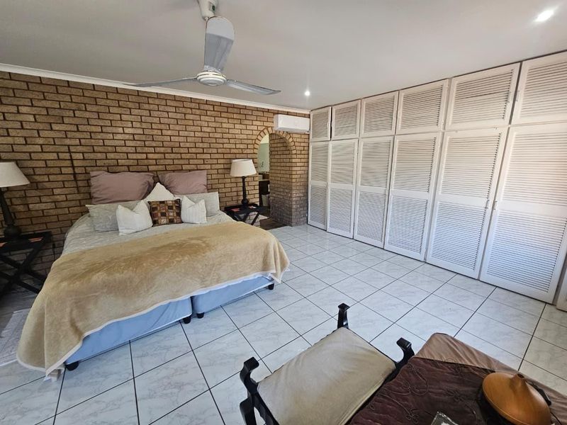 Spacious bedroom featuring an exposed brick feature wall, extensive built-in white louvered cupboards, tiled flooring, ceiling fan, and wall-mounted air conditioning unit.