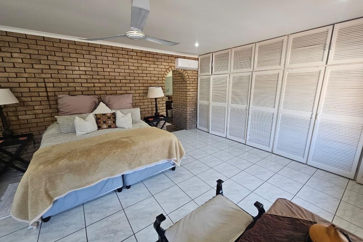 Spacious bedroom featuring an exposed brick feature wall, extensive built-in white louvered cupboards, tiled flooring, ceiling fan, and wall-mounted air conditioning unit.