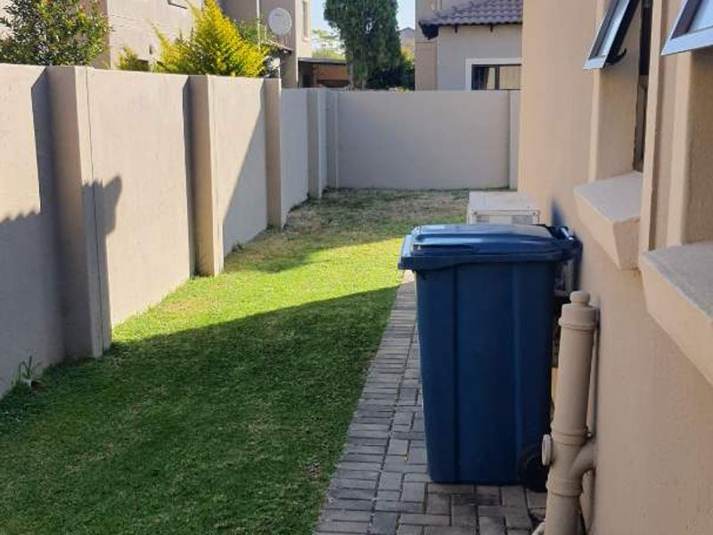 A side garden area featuring a paved walkway, lawn, and boundary wall, providing access along the side of the townhouse.