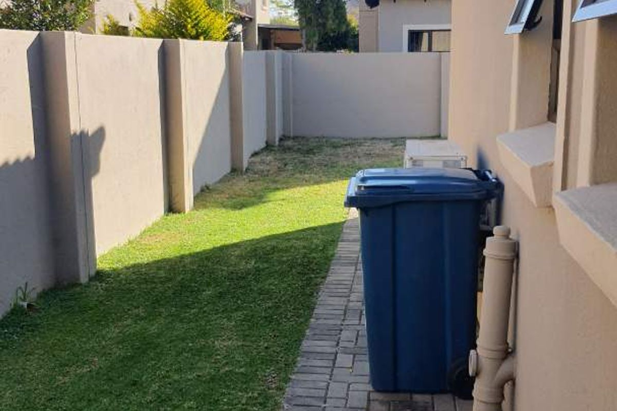 A side garden area featuring a paved walkway, lawn, and boundary wall, providing access along the side of the townhouse.