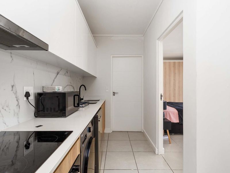 Modern galley-style kitchen featuring sleek white cabinetry, marble-look splashback, integrated electric hob, and tiled flooring, with direct access to the adjacent bedroom.