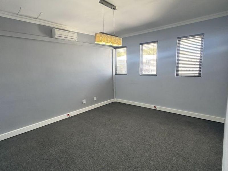 A clean, empty office space featuring grey carpeting, neutral walls, split-system air conditioning, and multiple windows with fitted venetian blinds.