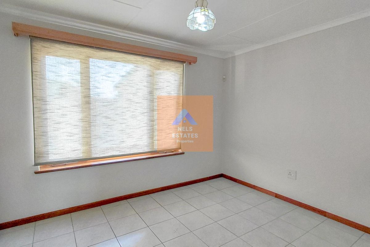 A bright, empty bedroom featuring tiled flooring, built-in cupboards, and a large window with a fitted roller blind.